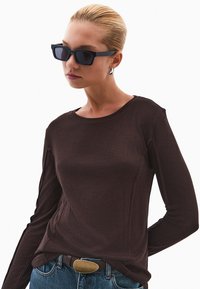 Donkerbruine top met lange mouwen, gemaakt van een zachte stof en met een ronde halslijn. Gecombineerd met blauwe jeans en een bruine riem met een ronde gesp.