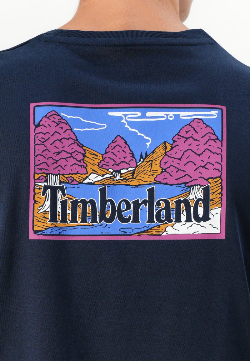Kolorowy krajobraz graficzny z różowymi drzewami, niebieskim niebem, górami i napisem "Timberland" na granatowej koszulce z tyłu.