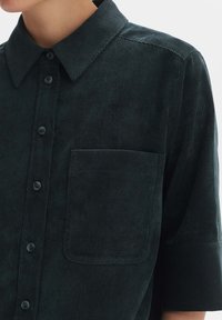 Dark green corduroy shirt with short sleeves, buttoned front, and a chest pocket; features textured ribbed fabric and a classic collar.