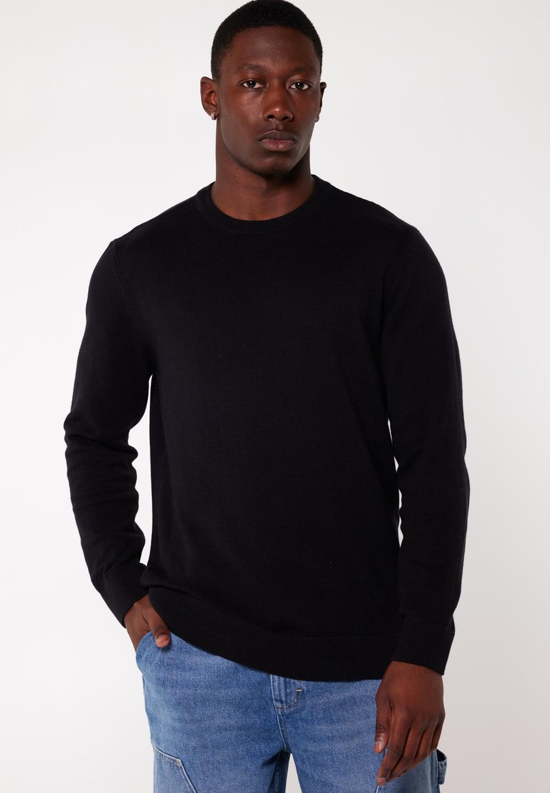 Black knit sweater with a round neckline, long sleeves, and ribbed cuffs and hem, worn over blue denim jeans. Textured fabric visible.