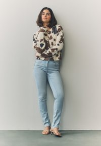 Pletena vestu u krem boji s smeđim i crnim mrljama poput krave, uparena sa svijetloplavim uskim trapericama i minimalističkim bež sandalama. Glatke teksture.