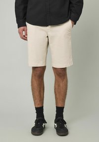 Shorts en coton beige avec poches latérales, devant plat et coupe droite. Portés avec une chemise noire à boutons et des baskets noires avec des accents blancs.