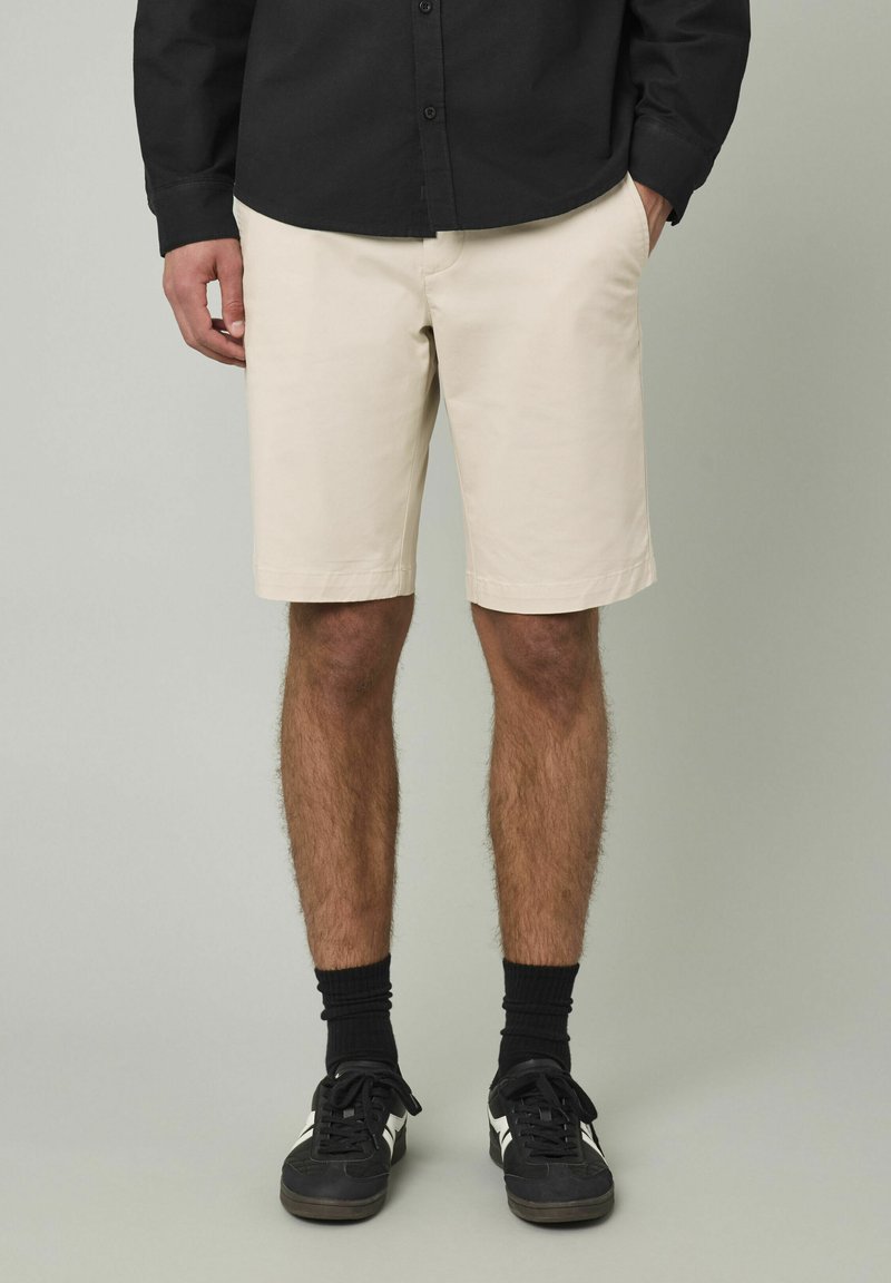 Shorts en coton beige avec poches latérales, devant plat et coupe droite. Portés avec une chemise noire à boutons et des baskets noires avec des accents blancs.