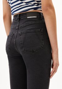 Henkilö, joka pukeutunut mustiin korkeavyötäröisiin farkkuihin, joissa on takataskut, ja raidallinen sinivalkoinen lyhyt paita, käsi lepää sivulla.