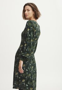 Dunkelgrünes Kleid aus weichem Stoff mit einem floralen Muster in Weiß und Gelb. Verfügt über lange, gepuffte Ärmel und einen lockeren Sitz.