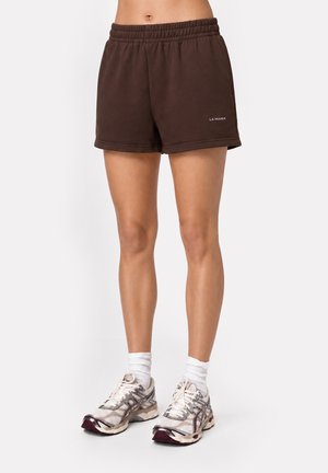 Kobieta nosząca brązowe elastyczne szorty z napisem "LA MANIA", białe skarpetki i białe buty do biegania, stojąca na białym tle.