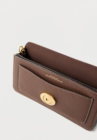 Καφέ πορτοφόλι από δερματίνη με υφή, με χρυσό στρογγυλό κλείσιμο και εσωτερική επένδυση από σουέντ, μερικώς ανοιχτό για να φαίνεται το εσωτερικό ετικέτα κειμένου.