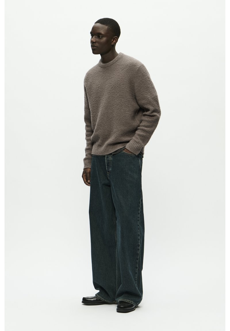 Pull en textured kaki, jeans oversized bleu foncé et chaussures noires. La tenue présente une coupe décontractée et des contrastes de couleurs subtils.