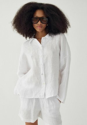 Žena s veľkým afro účesom, ktorá má na očiach veľké čierne slnečné okuliare, vo voľnej bielej lněnej košeli na gombíky a zladených šortkách, stojaca so založenými rukami v vreckách.