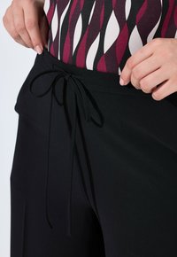 Zwarte broek met een gladde textuur en een verstelbare tailleband, gecombineerd met een gedessineerde top met golvende kastanjebruine en witte patronen.