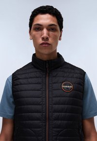 Fiatal férfi rövid, göndör hajjal, fekete, steppelt mellényt visel kék rövid ujjú ing fölött, semleges arckifejezéssel, egyenesen előre néz.