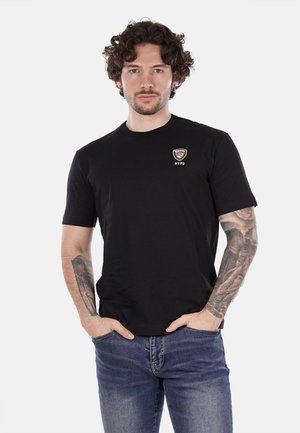 Maglietta nera a maniche corte realizzata in morbido cotone, con un piccolo logo colorato NYPD sul lato sinistro del petto; il modello indossa jeans blu.