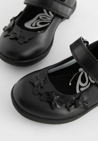 Chaussures noires en cuir de type Mary Jane avec une texture lisse, ornées de trois accents en forme de papillon sur le bout et d'une bride ajustable.