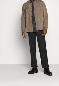 Camisa marrón de botones con dos bolsillos en el pecho sobre una camiseta negra con patrón de camuflaje, combinada con pantalones negros ajustados y botas negras hasta el tobillo.
