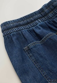 Primer plano de pantalones de mezclilla azul con cintura elástica y bolsillo trasero visible con costuras amarillas sobre un fondo claro.