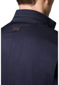 Granatowa kurtka z wyraźnym kołnierzem, minimalnymi detalami szycia i brązową skórzaną naszywką z logo z tyłu. Gładka, matowa faktura materiału.