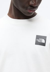 Άσπρο βαμβακερό φούτερ με τετράγωνο μαύρο λογότυπο με λευκά γράμματα "The North Face" στην αριστερή πλευρά του στήθους.