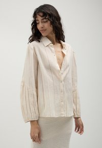 Woman in a cream-colored, long-sleeve button-up blouse with subtle stripes, paired with a textured beige skirt, looking down and to the side.