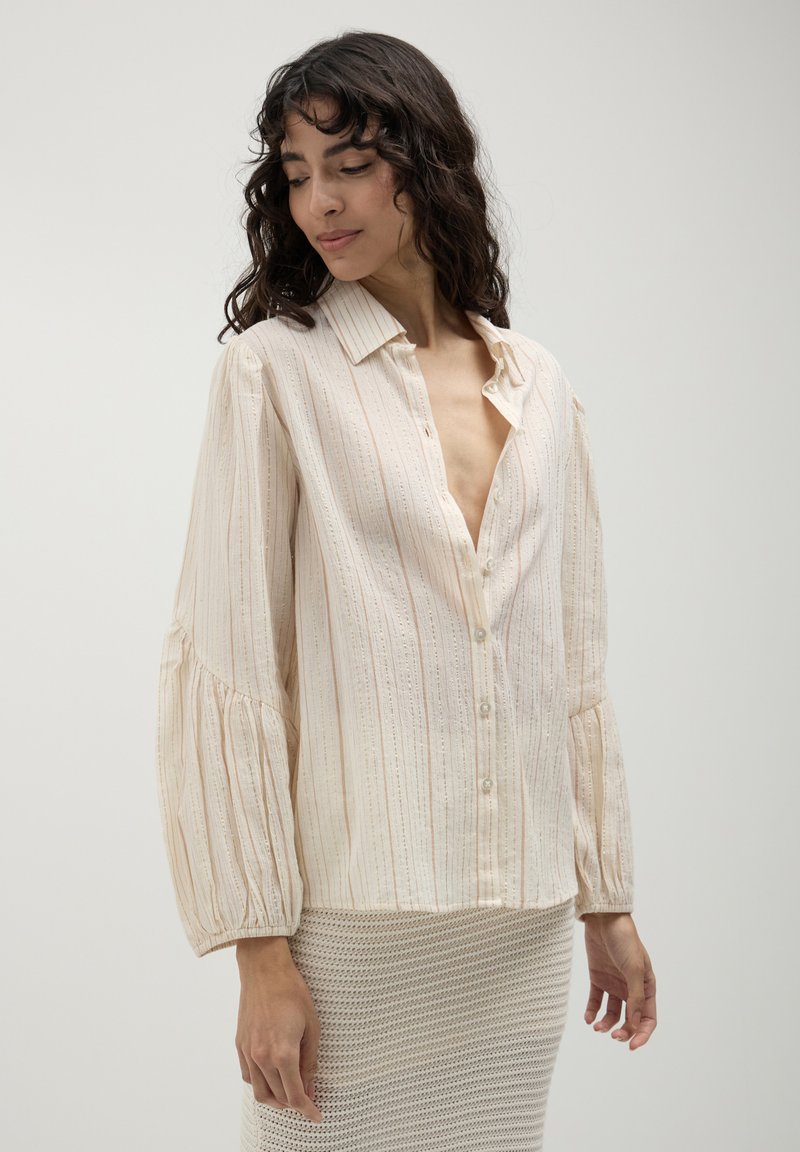 Woman in a cream-colored, long-sleeve button-up blouse with subtle stripes, paired with a textured beige skirt, looking down and to the side.