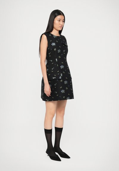 Fekete virágmintás ruha, testhez álló szabású, rövid ujjú, elülső zsebbel. Térdig érő fekete zoknikkal és hegyes orrú fekete cipőkkel párosítva.