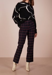 Pull noir avec motif abstrait blanc, associé à un pantalon à carreaux bleu marine avec des lignes orange. Le mannequin porte des bottines beige.