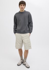 Grauer Rundhalspullover mit gerippten Bündchen, khakifarbene Cargo-Shorts mit Seitentaschen und grauen sowie beige Sneakers, stehend vor einem schlichten Hintergrund.