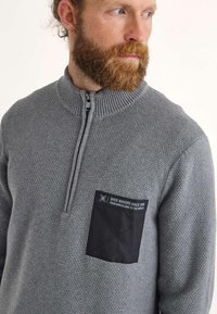 Pullover grigio testurizzato con colletto a mezza zip, dotato di una tasca a toppa nera sul petto con testo bianco e un design quadrato.