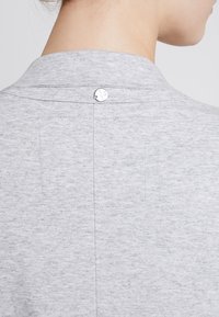 Harmaa neulepaita, jossa on kaulus, pehmeä rakenne ja koristeellinen nepparikiinnitys takana. Pienet kirjontadetaljit näkyvissä.