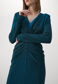 Teal glittrande klänning med långa ärmar, V-halsning och tvistad framsidesdetalj. Tillverkad av skimrande tyg som faller snyggt.