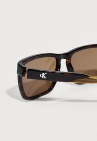 Okulary przeciwsłoneczne w kolorze żółwiowej skorupy z brązowymi soczewkami, kanciastą oprawą i srebrnym logotypem na zauszniku. Gładki plastikowy materiał i polerowane wykończenie.