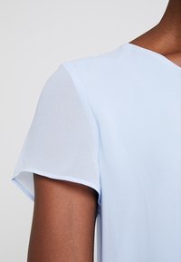 Light blue blouse featuring short, sheer sleeves and a v-neckline, crafted from a smooth fabric for a lightweight appearance.