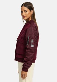 Burgundy bomber jacket with a textured front, smooth sleeves, ribbed cuffs, and zippered pocket on the left arm featuring patterned detailing.
