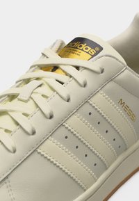 Κοντινό πλάνο λευκού αθλητικού παπουτσιού Adidas με κρεμ κορδόνια, χρυσό λογότυπο Adidas στη γλώσσα και χρυσό κείμενο "Messi" στο πλάι.