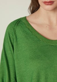 Zielony sweter z dzianiny o szerokim dekolcie, cechujący się luźnym krojem i miękką fakturą. Szczegóły obejmują prążkowane krawędzie i lekką tkaninę.