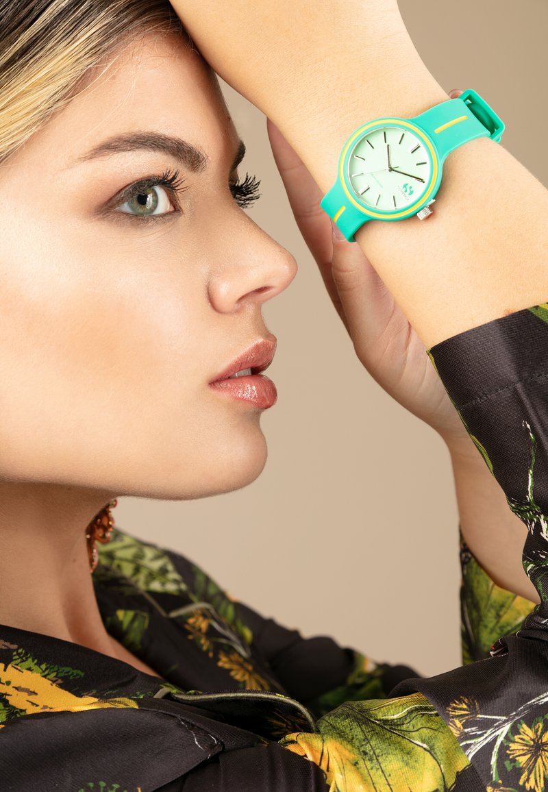 Reloj turquesa con una esfera verde redonda, manecillas finas, correa ajustable y acentos amarillos contrastantes; llevado en una muñeca contra una camisa estampada.