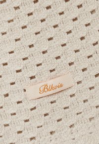 Tissu crocheté de couleur crème avec une étiquette rectangulaire beige cousue dessus, présentant un texte cursif orange "Blkvis" au centre.