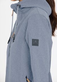 Light grey, textured jacket with a zippered hood, drawstring, tan accents, and a black logo patch on the left arm. Features zippered pockets.