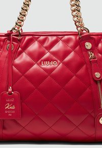 Rote gesteppte Handtasche aus glattem Leder mit goldfarbenen Elementen, einer Kettenhandhabe und einem Anhänger mit der Aufschrift "LIUJO Kate Re-Edition 2012."