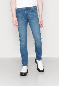 Person som bär slim-fit blå jeans och vita kraftiga stövlar, stående mot en enkel ljus bakgrund.