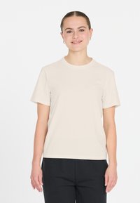 Beige bomull t-shirt med kort ärm och rundad halsringning, med diskret textdetalj på framsidan. Matchad med mörka byxor.