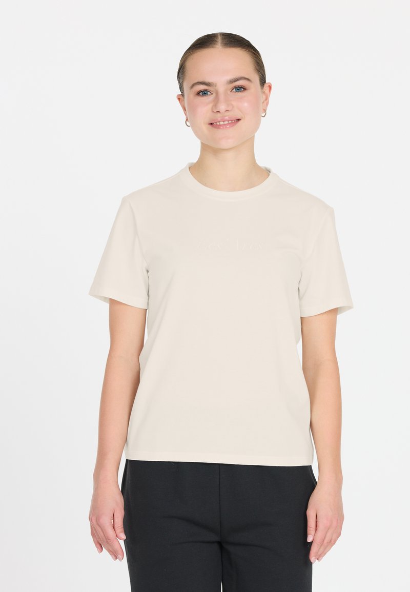 Beige bomull t-shirt med kort ärm och rundad halsringning, med diskret textdetalj på framsidan. Matchad med mörka byxor.