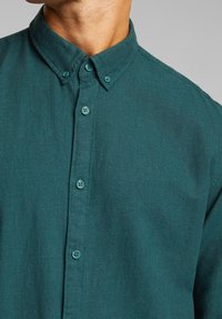 Camisa de botones azul verdoso con cuello, que presenta una tela texturizada y botones a juego en la parte delantera, sin patrones adicionales.