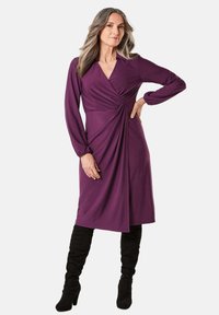 Lila Wickeldress mit langen Ärmeln, das ein gedrehtes Vorderdesign, eine weiche Textur und einen knielangen Saum aufweist, kombiniert mit schwarzen knielangen Stiefeln.