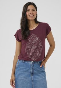 T-shirt bordeaux à manches courtes et col rond, avec un motif graphique léger détaillé. Associé à une jupe en denim bleue.