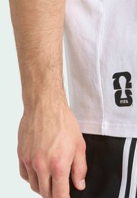 Bijela pamučna majica s crnim FIFA logotipom na donjem lijevom dijelu, u kombinaciji s crnim sportskim šortsevima s bijelim trakama.