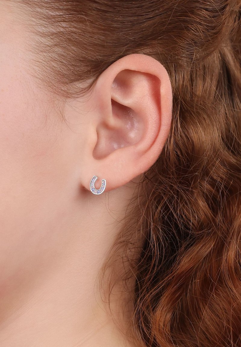 Pendientes en forma de herradura plateados con pequeñas piedras preciosas transparentes, sobre un tono de piel claro y peinados con cabello castaño suelto y rizado.
