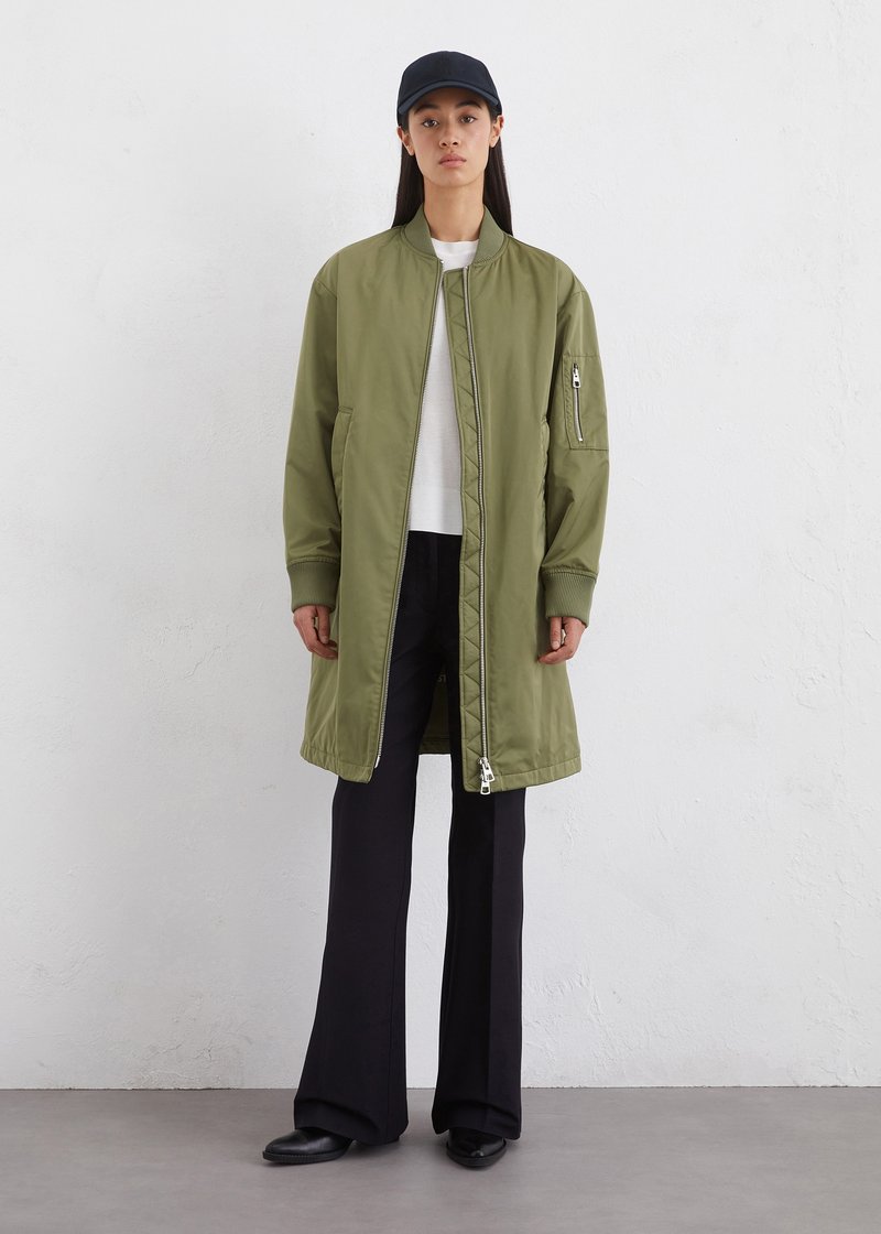 Olivgrüne Bomberjacke mit Frontreißverschluss, Seitentaschen und gerippten Bündchen, getragen über einem weißen Oberteil und schwarzen Schlaghosen. Eine schwarze Kappe rundet den Look ab.