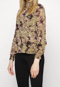 Blumige Bluse aus transparenter Stoff, mit hohem Kragen, langen Ärmeln mit gerafften Manschetten und einer Mischung aus grünen, lila und schwarzen Farbtönen.