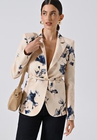 Beige blazer met marineblauw bloemenpatroon, reverskraag, ceintuur in de taille en twee voorkeepockets, gecombineerd met donkere jeans en een lichte handtas.