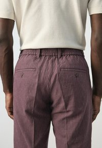 Hombre con pantalones morados de rayas finas y una camisa crema de manga corta, mostrado de espaldas, de pie contra un fondo liso.